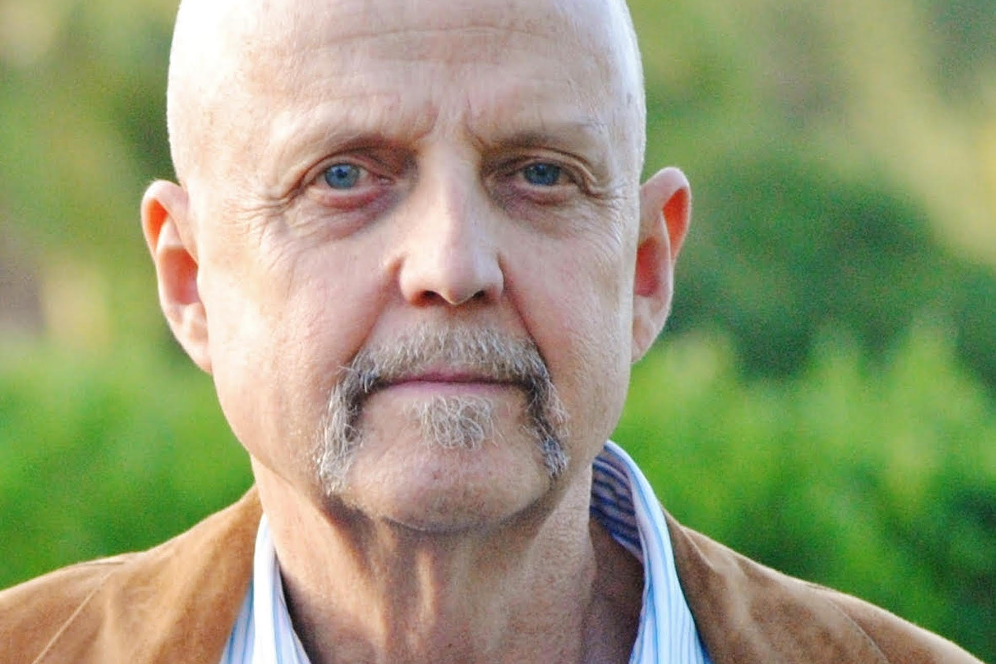Headshot of poet Ronald Koertge, a bald white male wearing a brown suede jacket
